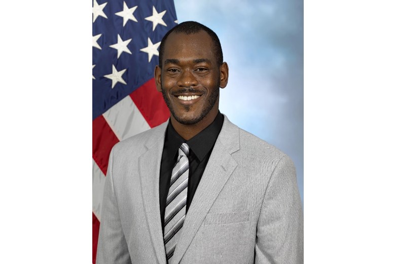 A man in a suit stands in front of an American flag.