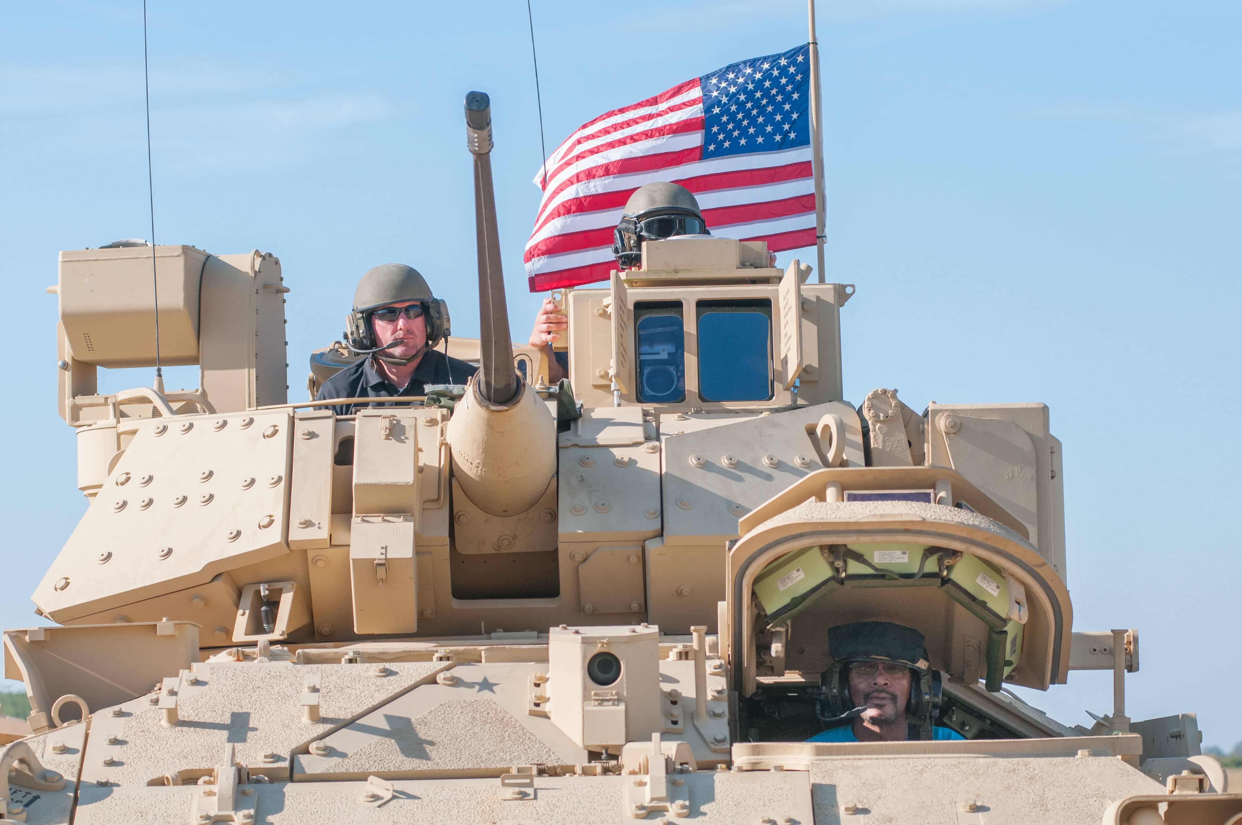 A civilian contractor and Soldier ride in a Bradley tank with an American flag in the background.