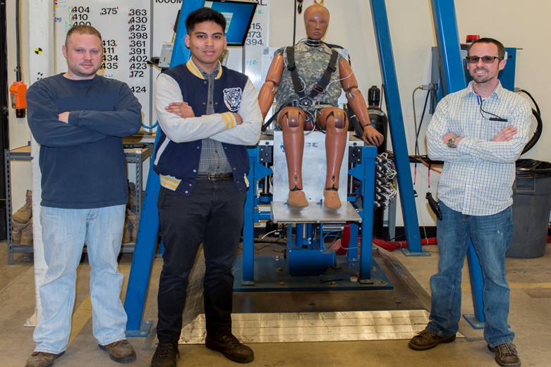 A group of men stand with arms crossed in front of a crash test dummy