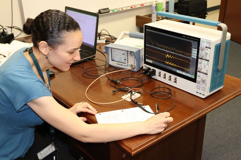 A woman takes notes on a notepad in front of an oscilloscope.