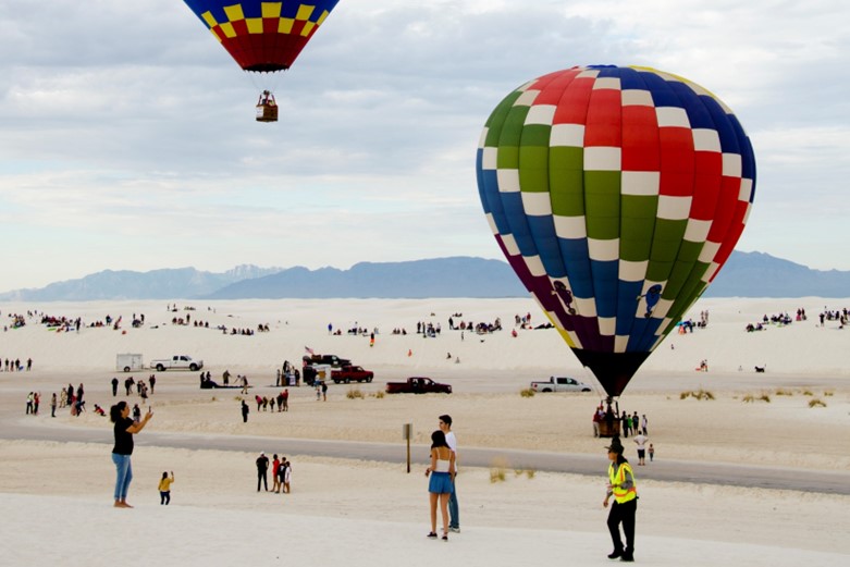 A desert scene with people on the ground and hot air balloons in the background