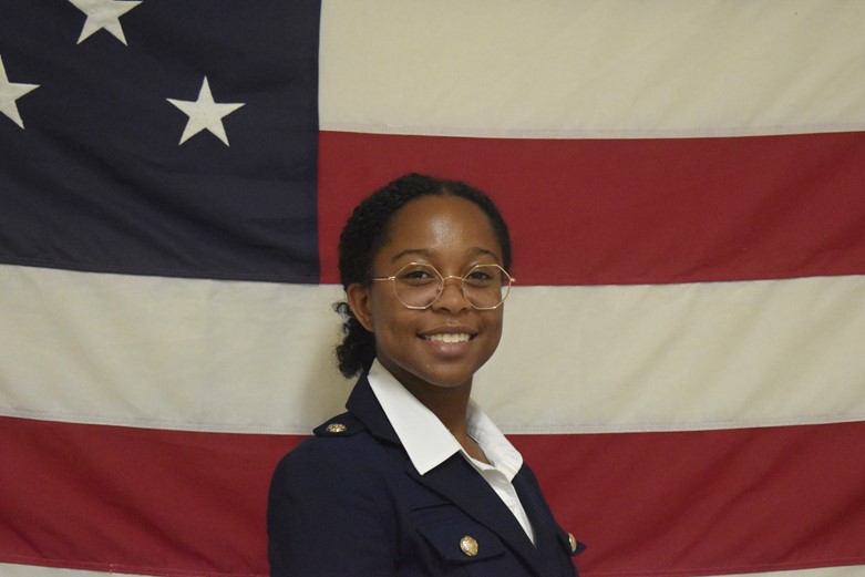 A woman in glasses stands in an Army dress uniform stands in front of a U.S. flag