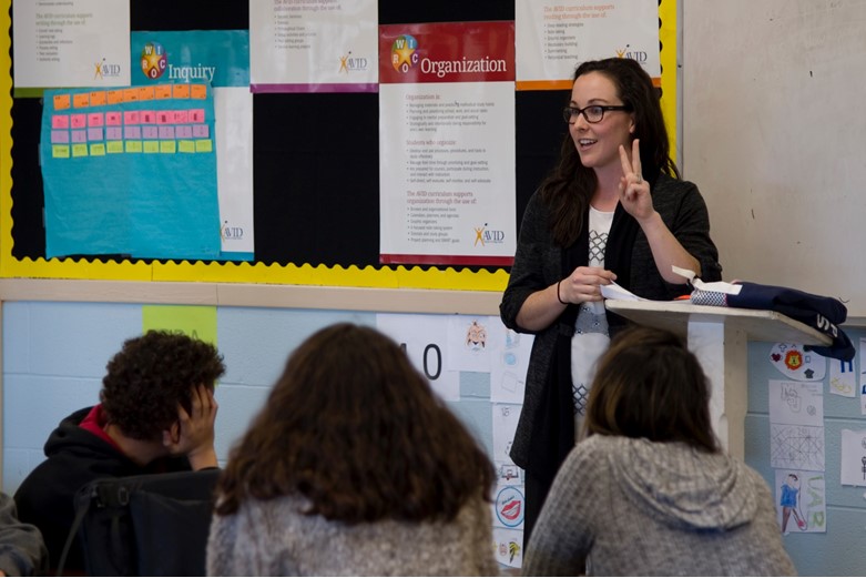 A woman presents information in the front of a class to students.